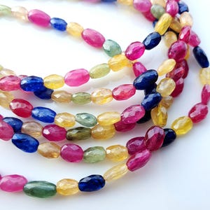 Könnte beinhalten: Eine Strähne ovaler Edelsteinperlen in verschiedenen Farben, darunter Blau, Grün, Gelb und Pink. Die Perlen sind facettiert und haben eine polierte Oberfläche.