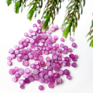 May include: A pile of small, faceted pink ruby gemstones. The rubies are hexagonal in shape and have a shiny, polished finish.