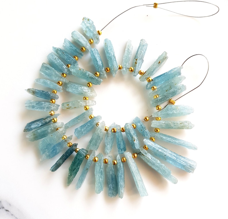 May include: A strand of light blue aquamarine crystal beads with gold spacers. The beads are long and narrow, resembling shards of crystal. The strand is strung on a wire and is ready to be used for jewellery making.