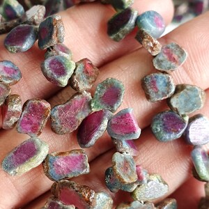 May include: A strand of ruby zoisite gemstone beads, featuring a mix of green, pink, and brown hues. The beads are irregularly shaped and have a rough, natural finish.