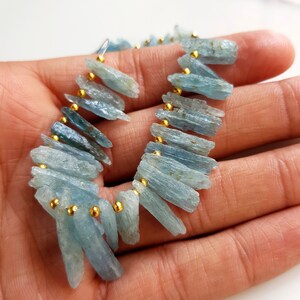 May include: A strand of light blue aquamarine gemstone beads with gold spacers. The beads are long and narrow, with a rough, natural finish.