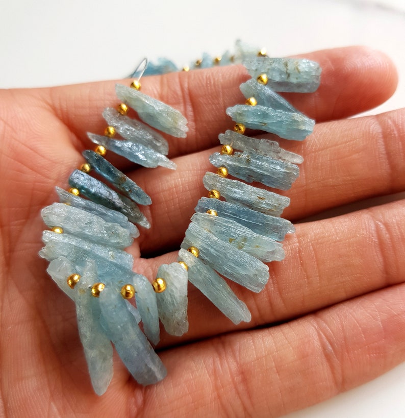 May include: A strand of light blue aquamarine gemstone beads with gold spacers. The beads are long and narrow, with a rough, natural finish.