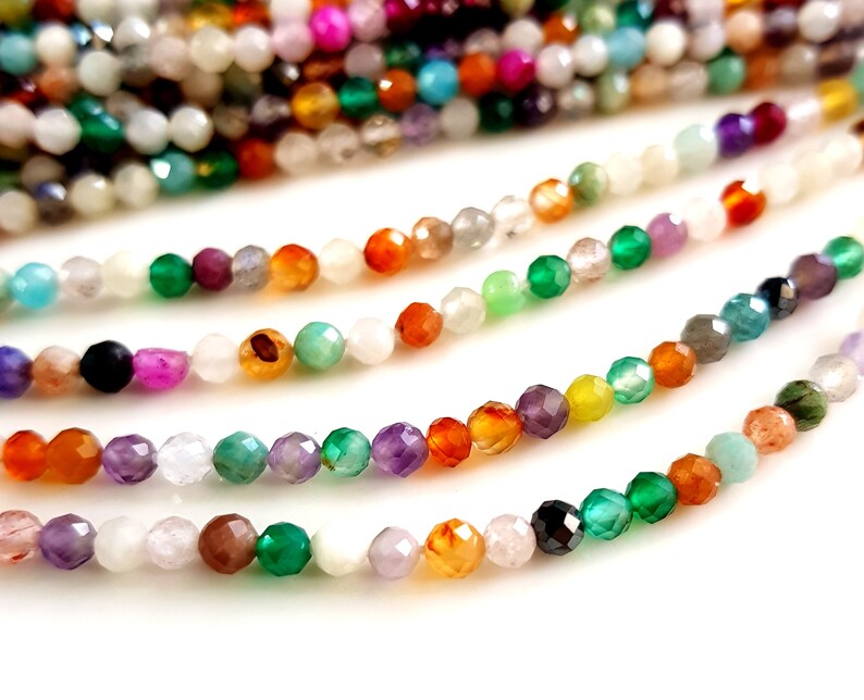 May include: Close-up of several strands of faceted gemstone beads in a variety of colors, including green, orange, purple, and white. The beads are strung together, creating a colorful and textured display. Ideal for jewelry making.