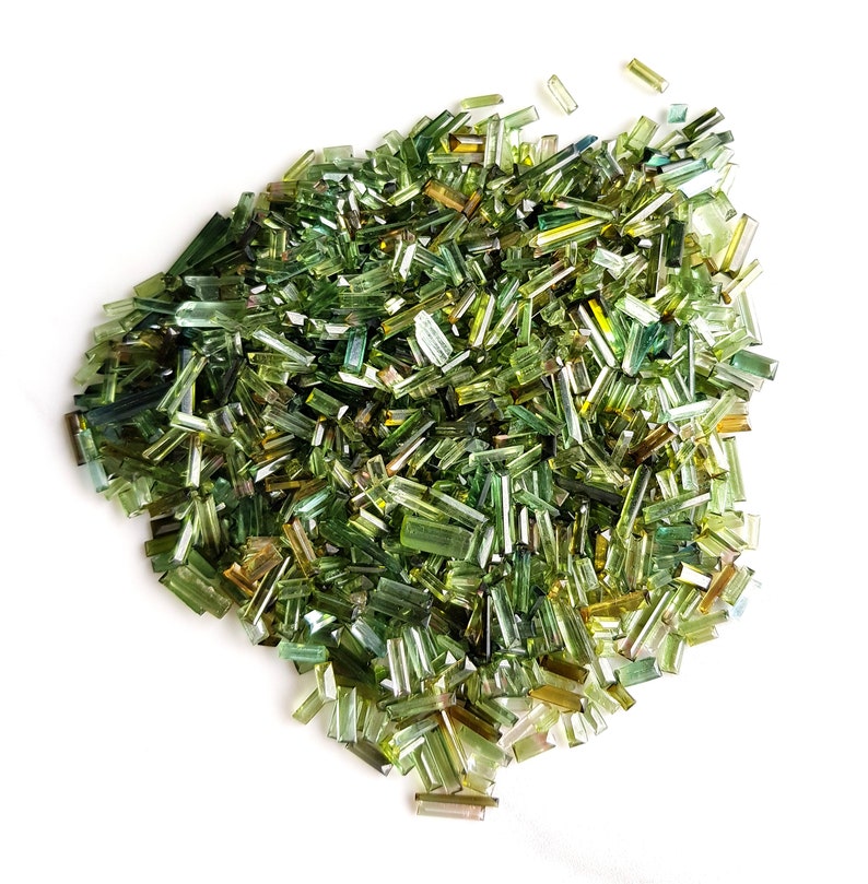 May include: A large pile of green and brown gemstone chips. The chips are long and thin, and they are scattered randomly on a white background.