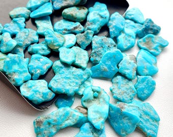May include: A collection of raw turquoise gemstones in various shapes and sizes. The stones are a vibrant turquoise blue with natural matrix patterns, displayed in a small black scoop. The gemstones are scattered across a white surface.