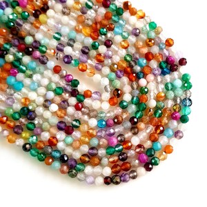 May include: A collection of faceted gemstone beads in various colors, including green, orange, purple, and white. The beads are small and round, with a sparkling, polished finish. The beads are scattered on a white surface.