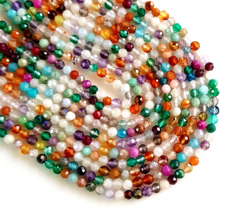 May include: A collection of faceted gemstone beads in various colors, including green, orange, purple, and white. The beads are small and round, with a sparkling, polished finish. The beads are scattered on a white surface.