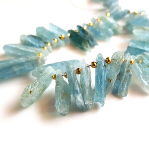 May include: A strand of aquamarine gemstone beads, each bead is a long, thin, and slightly irregular shard. The beads are strung on a wire with gold-coloured spacers between each bead.