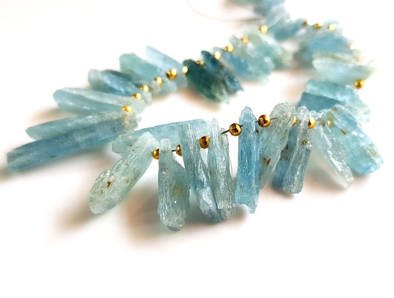 May include: A strand of aquamarine gemstone beads, each bead is a long, thin, and slightly irregular shard. The beads are strung on a wire with gold-coloured spacers between each bead.