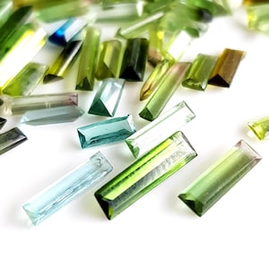 May include: Assortment of green, blue, and clear faceted gemstone beads in a rectangular shape. The beads are scattered on a white surface.
