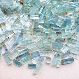 Pode incluir: Uma coleção de cristais de água-marinha brutos, exibindo uma variedade de tons azul claro. Os cristais variam em tamanho e forma, com texturas e inclusões naturais visíveis. As pedras preciosas estão espalhadas.
