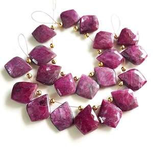 Puede incluir: Una hebra de cuentas de rubí facetadas, cada cuenta es de color rojo intenso con una forma ligeramente irregular. Las cuentas están enhebradas en un alambre fino con separadores de color dorado entre cada cuenta.