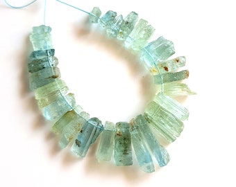 May include: A strand of light blue aquamarine gemstone beads, each bead is a long, thin, and faceted crystal. The beads are strung on a thin blue cord and are arranged in a circle.
