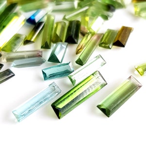 May include: Assortment of green, blue, and clear faceted gemstone beads in a rectangular shape. The beads are scattered on a white surface.