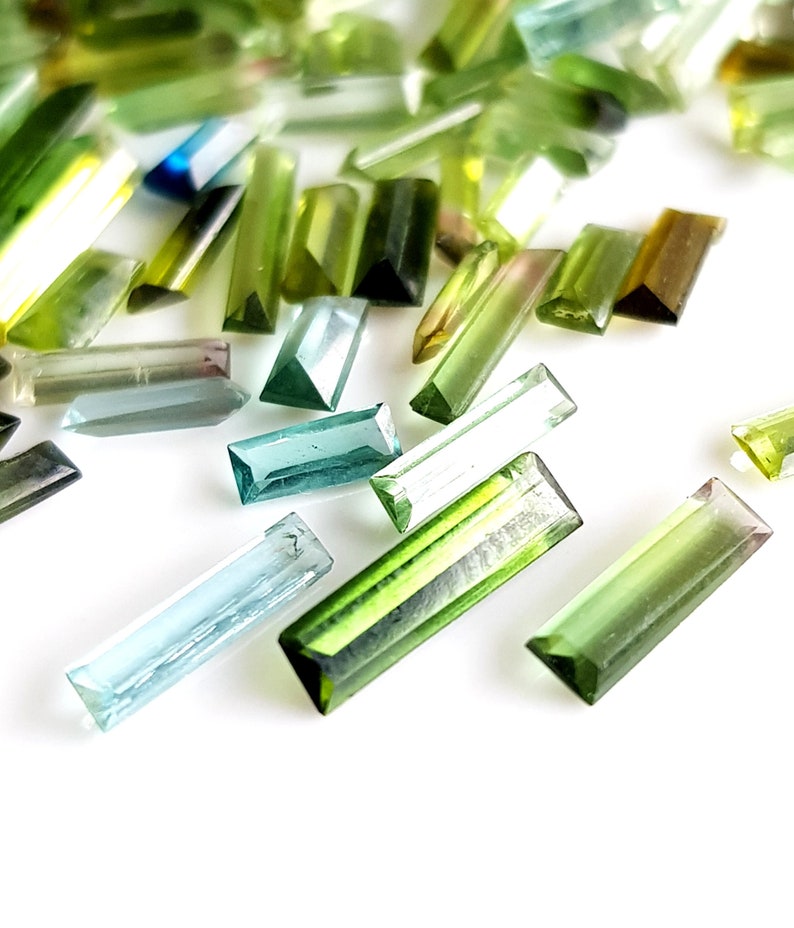 May include: Assortment of green, blue, and clear faceted gemstone beads in a rectangular shape. The beads are scattered on a white surface.