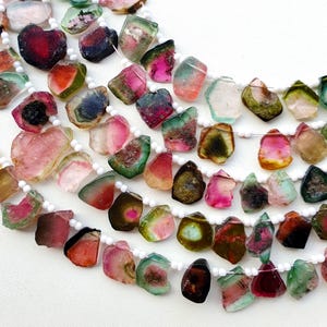 Può includere: Fili di perline di tormalina anguria di varie forme e dimensioni. Le perline mostrano un mix di colori, tra cui rosa, rosso, verde e trasparente, con perline distanziatrici bianche. Le pietre preziose sono infilate su un cordoncino bianco.