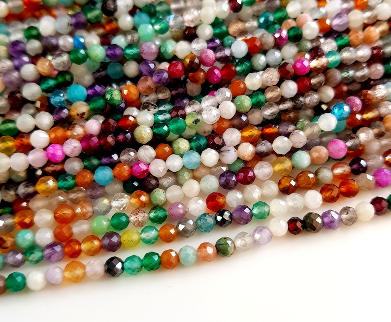 May include: A close-up shot of a collection of faceted gemstone beads in various colors. The beads are strung together, showcasing a diverse palette of hues including green, purple, orange, and white. These beads are suitable for jewelry making.