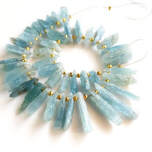 May include: A strand of light blue aquamarine gemstone beads with gold spacers. The beads are long and slender, resembling shards of crystal. The beads are strung on a clear thread.