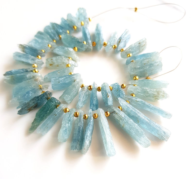 May include: A strand of light blue aquamarine gemstone beads with gold spacers. The beads are long and slender, resembling shards of crystal. The beads are strung on a clear thread.