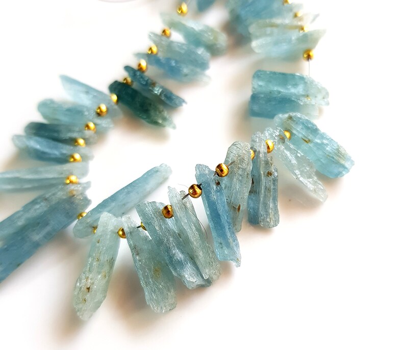 May include: A strand of aquamarine gemstone beads, each bead is a long, thin, and slightly irregular shape. The beads are strung on a wire with gold-coloured spacers between each bead.