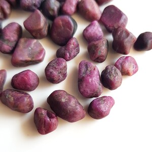 May include: A collection of small, rough, purple gemstones. The stones are various shapes and sizes, and have a natural, unpolished look.
