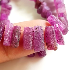 May include: A strand of purple rough gemstone beads. The beads are rectangular and have a rough, textured surface.