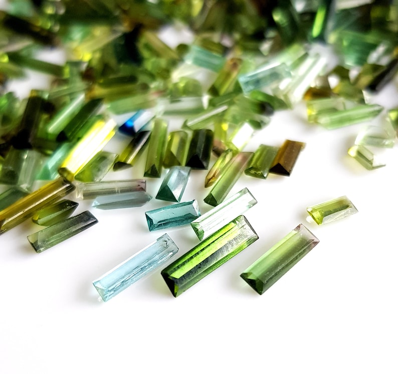 May include: A collection of green, blue, and clear faceted gemstone beads in a rectangular shape. The beads are scattered on a white surface.