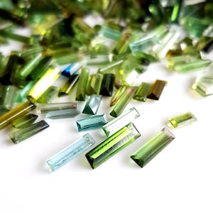 May include: A collection of green, blue, and clear faceted gemstone beads in a rectangular shape. The beads are scattered on a white surface.