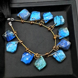 May include: A strand of blue labradorite gemstone beads strung on a gold wire with gold beads. The beads are faceted and have a shimmering iridescent effect.
