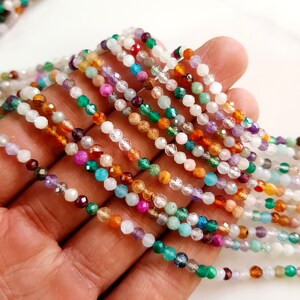 May include: Close-up of multiple strands of colorful gemstone beads. The beads are small and faceted, displaying a variety of colors including white, green, purple, orange, and red. The beads are strung together, creating a delicate and intricate design.
