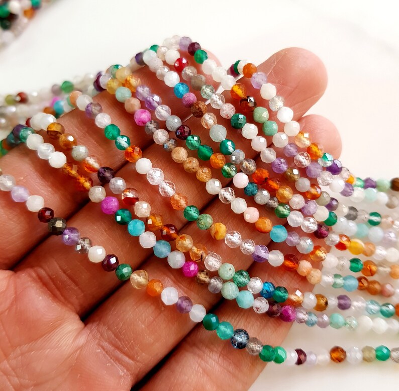 May include: Close-up of multiple strands of colorful gemstone beads. The beads are small and faceted, displaying a variety of colors including white, green, purple, orange, and red. The beads are strung together, creating a delicate and intricate design.