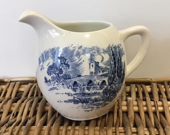 May include: A white ceramic milk jug with a blue and white landscape design. The jug has a rounded body and a handle.