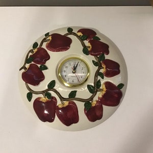 May include: White ceramic clock with a gold rim and black hands. The clock face is surrounded by a vine with red apples.