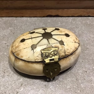 May include: A small, oval-shaped box made of bone with a brass latch and a geometric design on the top. The box is closed.