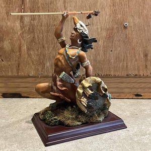May include: A detailed figurine of a Native American warrior in a kneeling pose, holding a spear aloft. The figure is adorned with a headdress, jewelry, and a shield. The base is a dark brown rectangular shape, and the overall color palette is earthy tones.