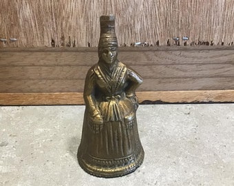 Op de afbeelding: Een gouden metalen bel in de vorm van een vrouw die een jurk en een hoed draagt. De bel heeft een gedetailleerd ontwerp en is van traditionele stijl.