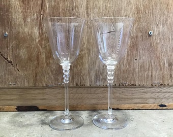May include: Two clear glass wine glasses with a stem and a round base. The glasses are empty and have a textured design.