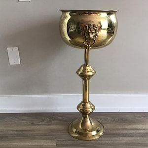 Puede incluir: Un alto soporte para plantas de metal dorado con un cuenco redondo y asas con cabeza de león. El soporte tiene una base decorativa y una columna alta y delgada.