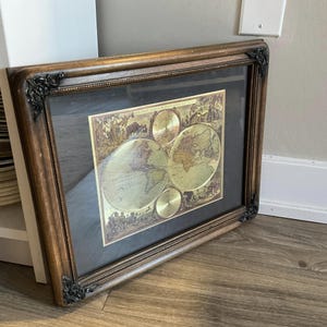 May include: A framed gold-leaf world map with a dark brown wooden frame. The map is surrounded by a decorative border and includes a compass rose and other details.