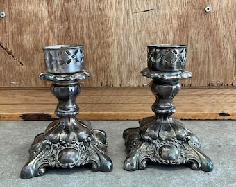 May include: A pair of silver candle holders with ornate designs and a cut-out pattern on the top. The candle holders have a square base with intricate details.
