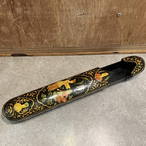 May include: A long, black, decorative box with a sliding lid. The box is adorned with gold and colorful floral and figure designs. The interior is black. The box is resting on a gray surface.