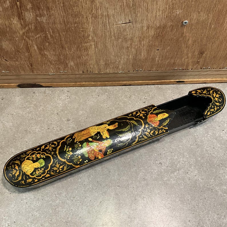 May include: A long, black, decorative box with a sliding lid. The box is adorned with gold and colorful floral and figure designs. The interior is black. The box is resting on a gray surface.