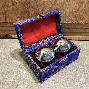 May include: A pair of shiny, silver metal exercise balls rest inside a blue, ornate box with a red interior. The box is open, revealing the balls and the red lining. The box has a gold clasp.