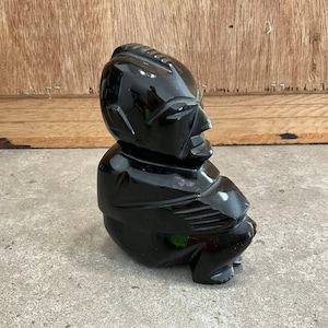 Pode incluir: Escultura preta brilhante de uma figura sentada com cabeça e corpo estilizados. A figura tem uma superfície lisa e polida, provavelmente esculpida em um material sólido. A escultura está sobre um fundo neutro.