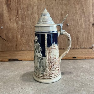 以下が含まれることがあります： ヒンジ付きの金属製の蓋が付いた、ヴィンテージのセラミックビールジョッキ。ジョッキには、女性と建物のレリーフ彫刻が施されており、青とクリーム色の配色です。ハンドルは湾曲しており、ジョッキの高さは約25cmです。