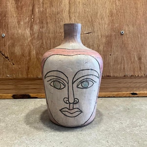 May include: A ceramic vase shaped like a face with pink, white, and green paint. The vase has a wide mouth and a narrow base.