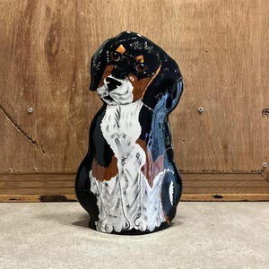 May include: A ceramic figurine of a black, brown, and white dog sitting with its head tilted to the side. The dog has a black nose and brown eyes.