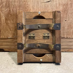 Puede incluir: Una caja de madera con dos bisagras de latón y un cierre de metal. La caja está cerrada y tiene un aspecto rústico.