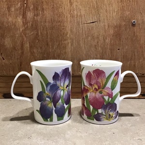 May include: Two white ceramic mugs with floral designs. One mug features purple and blue irises, while the other mug features pink and yellow irises.
