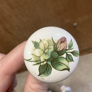May include: White ceramic cabinet knob with a floral design. The knob features a painted illustration of a pink and green rose with green leaves. The knob is round and has a slightly aged appearance.